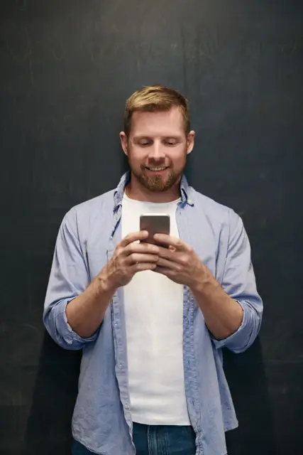 A cheerful man using a smartphone against a chalkboard indoors.